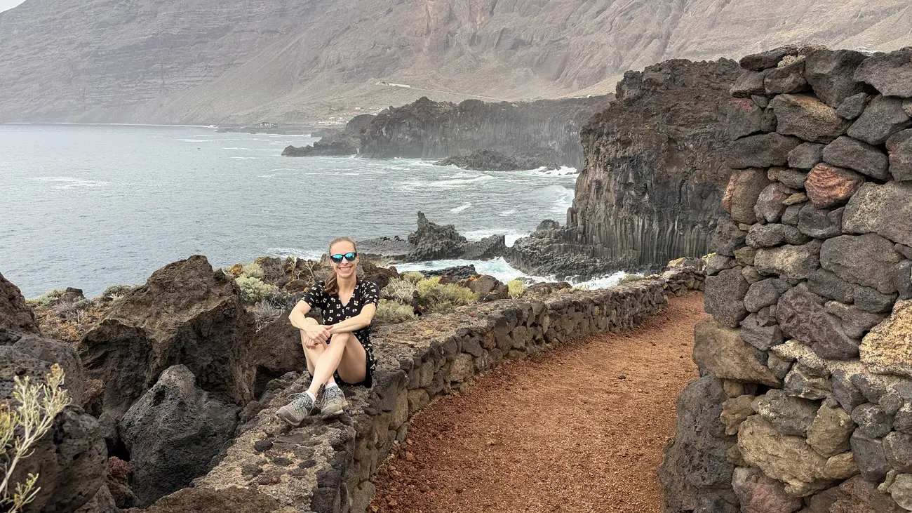 Mujer sentada junto al sendero litoral de Las Puntas con el paisaje volcánico de El Hierro al fondo